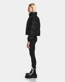 A woman stands in profile wearing the Rudsak OPRAH - 8124708 Black cropped down puffer with removable sleeves, black leggings, and black boots with white fur trim against a plain light gray background.