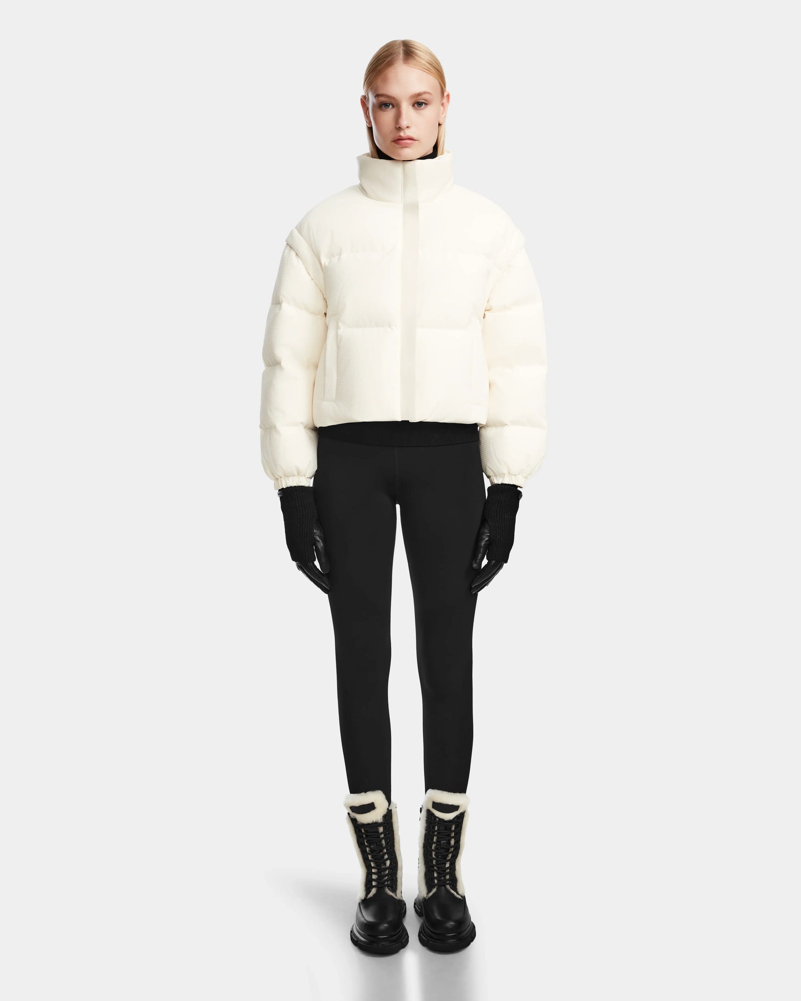 A woman stands facing forward wearing the Rudsak OPRAH - 8124708 Iceland womens cropped down puffer jacket with black gloves, leggings, and winter boots with white trim against a plain light gray background.