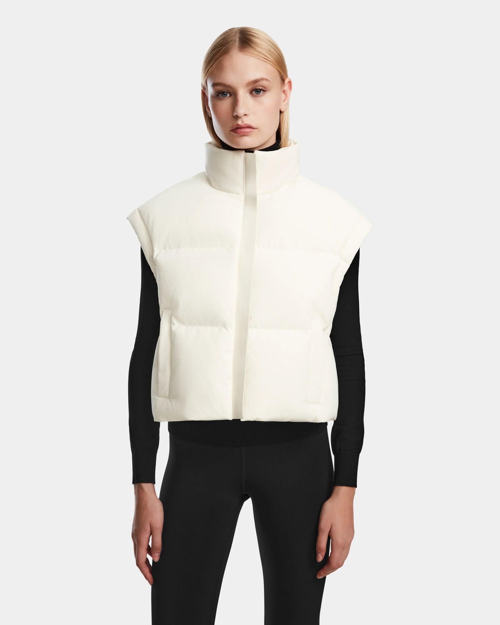 A woman with straight blonde hair wears the Rudsak OPRAH - 8124708 Iceland womens cropped down puffer, layered over a black long-sleeve shirt and black pants, standing against a plain light gray background.
