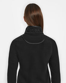 A person with straight brown hair wears the Rudsak ORCHID - 8524616 Black jacket, featuring vegan leather trims and a high collar with RUDSAK embossed on the back. The person faces away, against a white background.