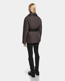 A person with straight blonde hair, dressed in the Rudsak PAOLA - 8124547 Grey RDS-certified down puffer jacket, black leggings, and black knee-high boots stands facing away against a plain light background.