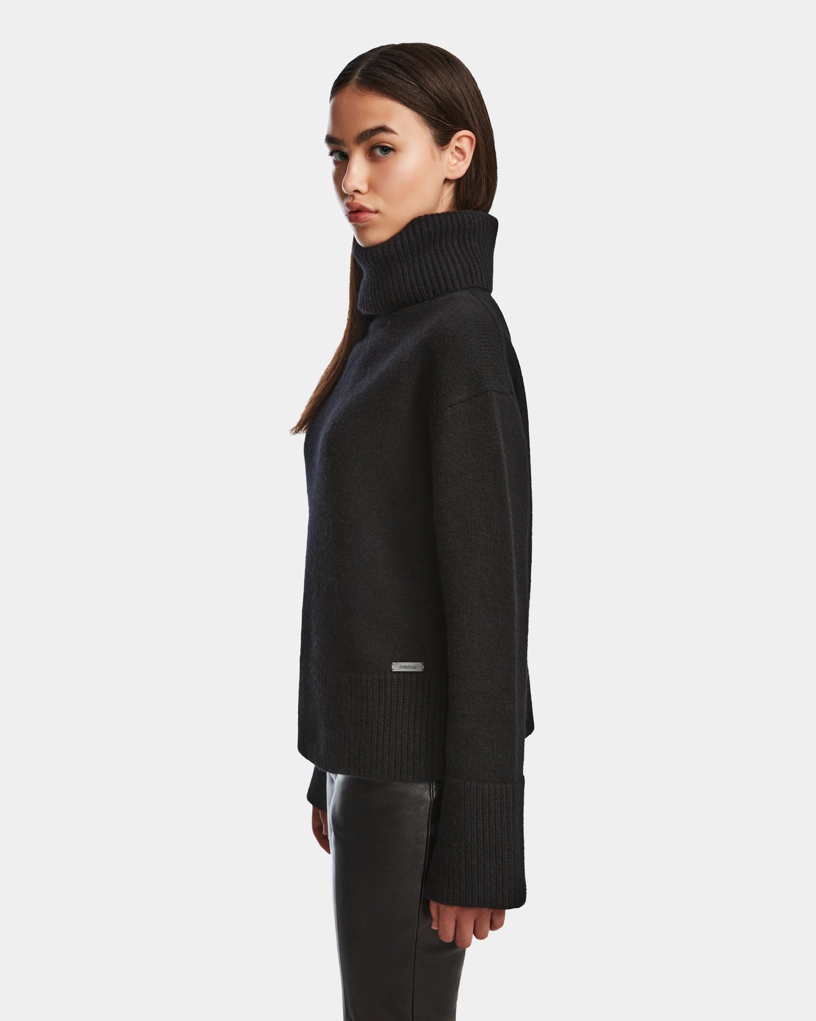 A woman with straight brown hair wears the Rudsak PARMI - 8524608 Black womens knit turtleneck sweater and black leather pants, standing in profile against a plain white background.