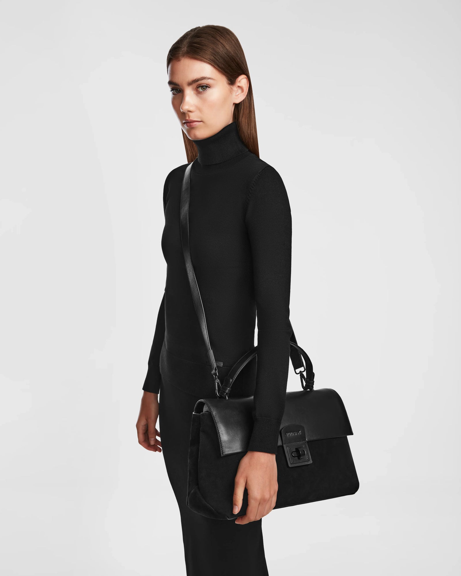 A woman in a black turtleneck dress stands sideways, holding the Rudsak ESMA - 8324519 Black handbag made from suede and leather with a removable shoulder strap and top handle, against a plain white background.