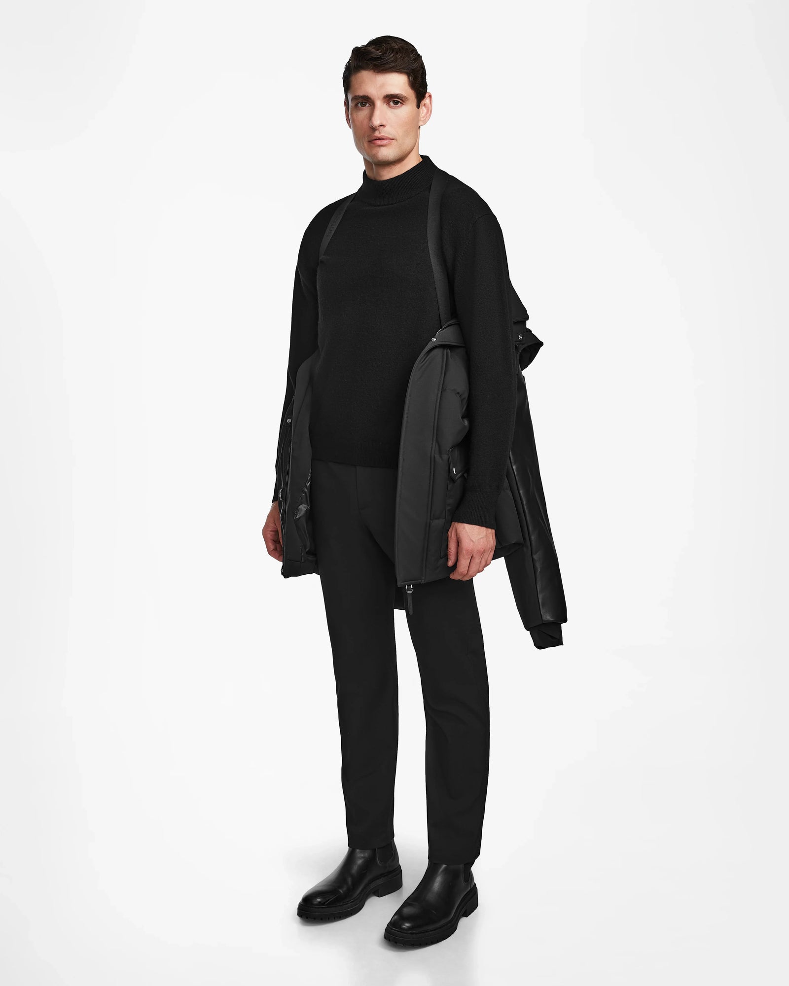 A man stands against a white background wearing a black turtleneck, pants, boots, and draping the Rudsak RICKY R - M123506 Black men's quilted down parka with recycled leather sleeves over his shoulders, looking straight ahead with a neutral expression.