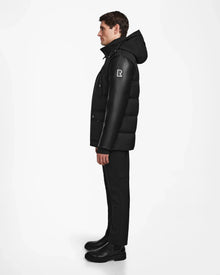 A man stands in profile wearing the Rudsak RICKY R - M123506 Black men's quilted down parka with recycled leather sleeves, paired with black pants and boots, against a plain white background.