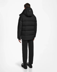 A person stands with their back to the camera, wearing Rudsak's RICKY R - M123506 Black men’s quilted down parka with recycled leather sleeves, black pants, and black shoes against a plain white background.