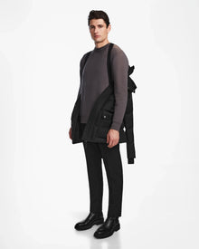 A man stands against a white background, wearing a gray sweater, black pants, black boots, and carrying the Rudsak RICKY - M123502 Black parka over one shoulder. He looks forward with a neutral expression.