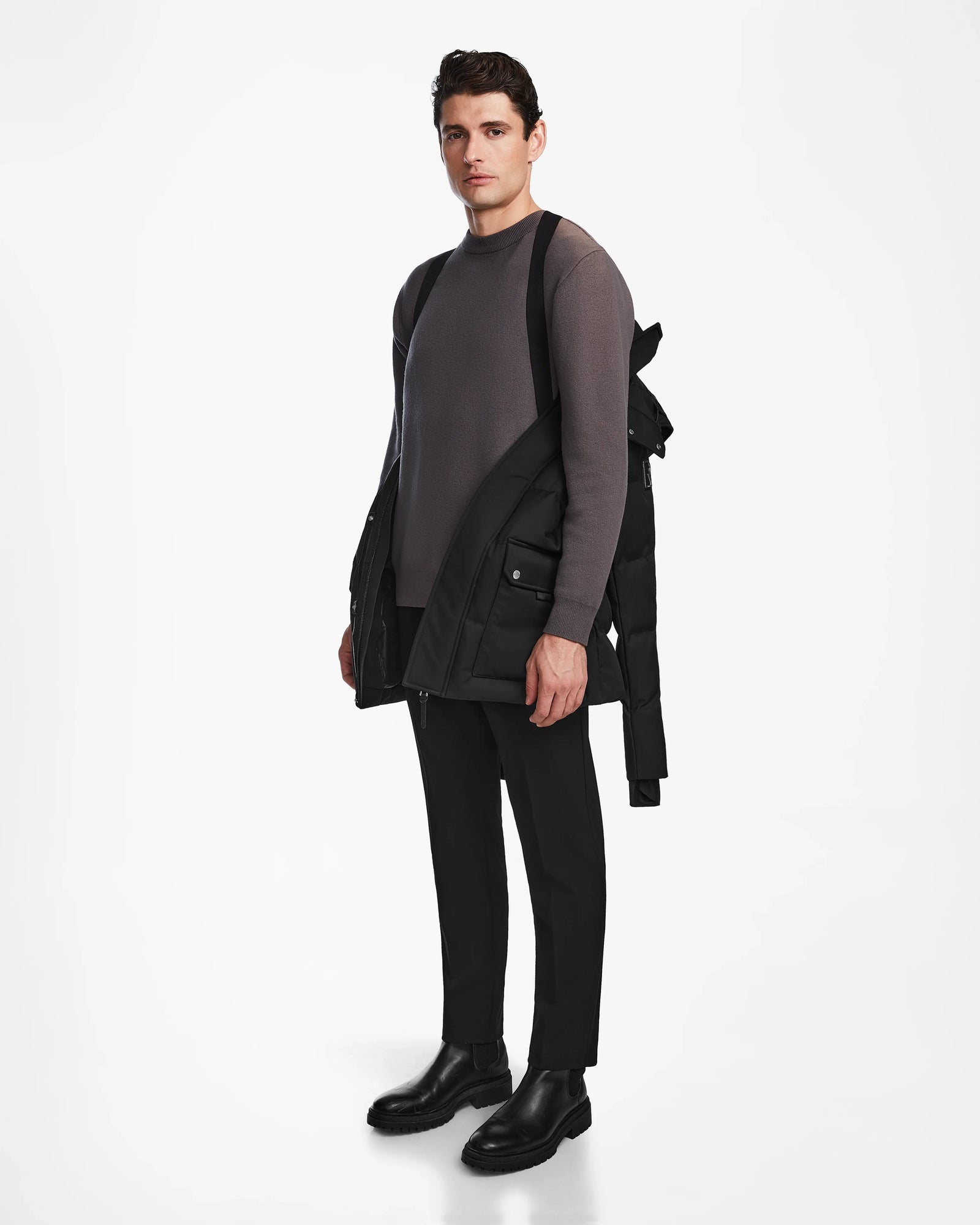 A man stands against a white background, wearing a gray sweater, black pants, black boots, and carrying the Rudsak RICKY - M123502 Black parka over one shoulder. He looks forward with a neutral expression.