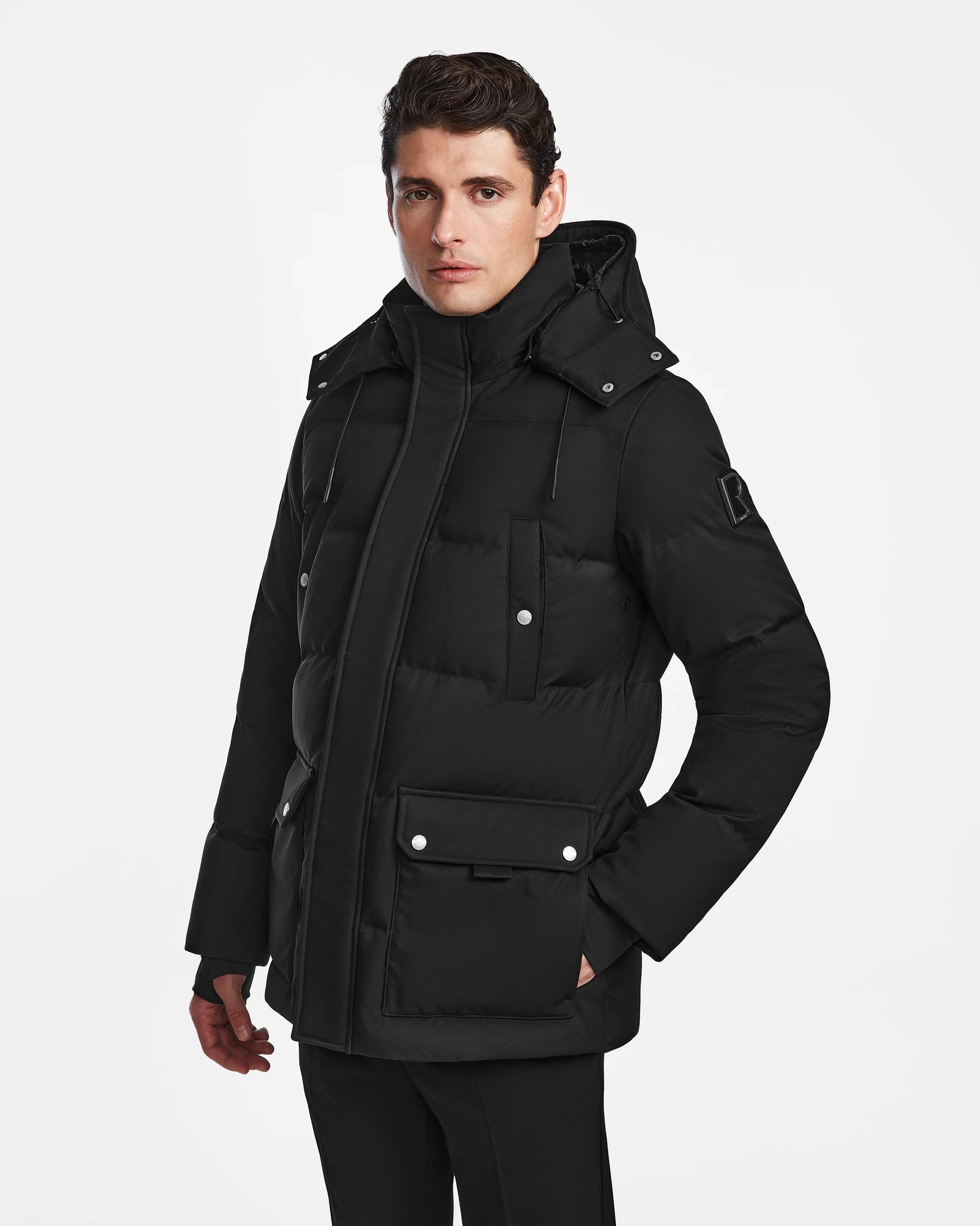 A man models the Rudsak RICKY - M123502 black puffer jacket with a hood, large front pockets, and button details, standing against a plain white background and looking slightly to the side.