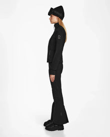 A woman stands in profile against a plain white background, wearing the Rudsak ROSALIE - 8124784 Black fitted ski jacket, flared pants, winter boots, and a knit beanie hat with RUDSAK written on it.