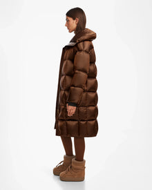 A woman stands in profile wearing the Rudsak SASH R - 8124543 Antique Bronze women’s long down puffer coat with a large collar, brown tights, and tan winter boots against a plain white background. The coat features RDS-certified down for warmth and comfort.