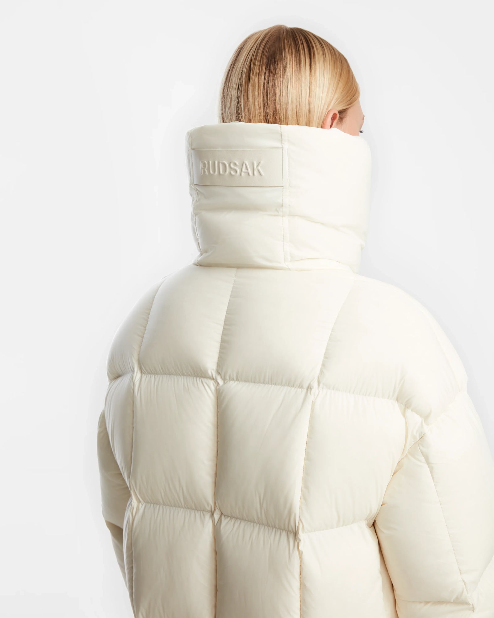 A person with straight blonde hair wears the off-white Rudsak SASH - 8124536 Iceland womens down puffer coat with quilted stitching and a high, branded collar. The background is plain white.