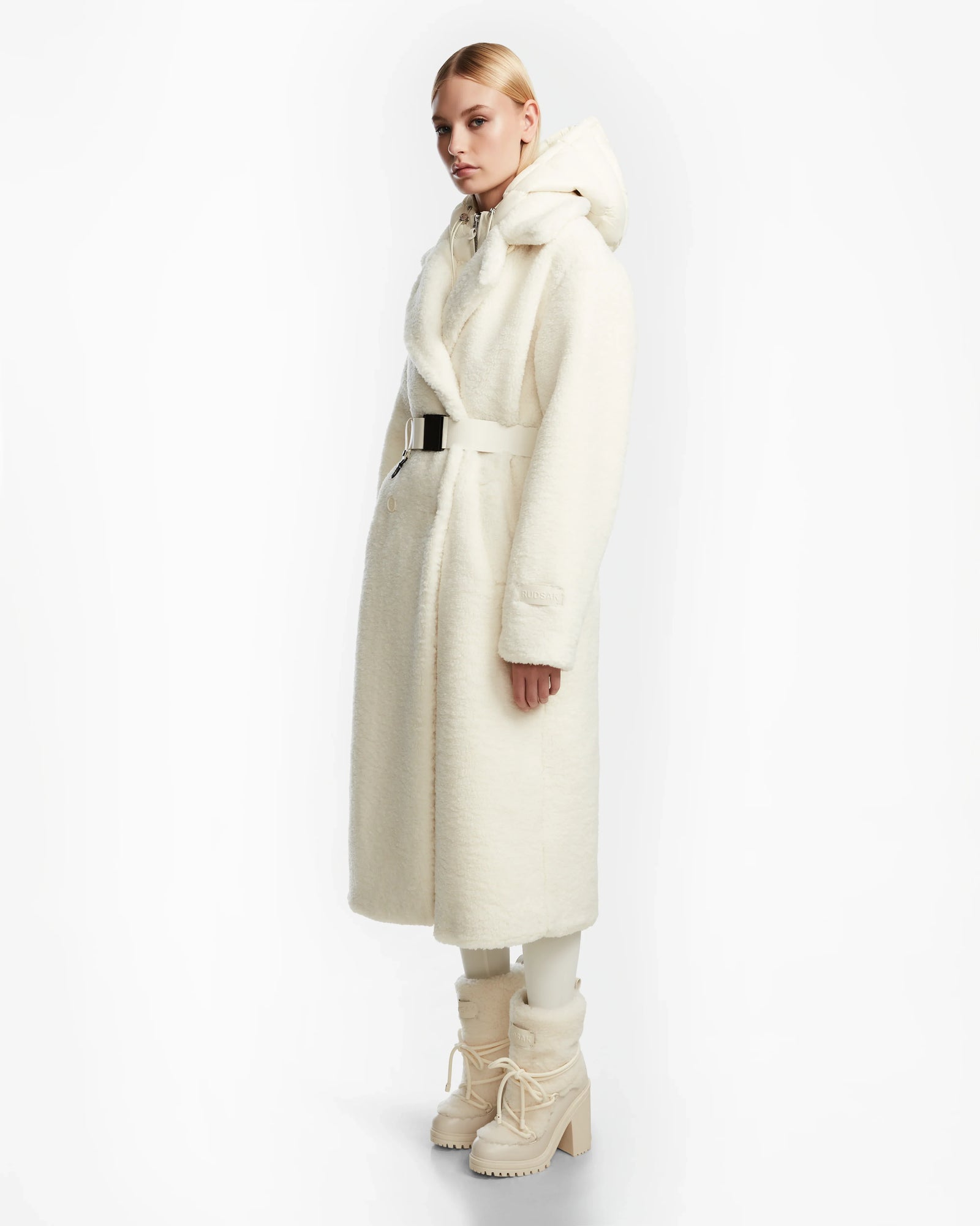 A person poses against a white background wearing the Rudsak TINA - 8123578 Iceland womens faux fur teddy trench coat with a belt, styled with matching boots and light-colored pants.