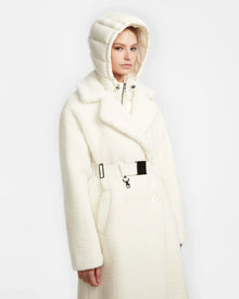 A woman models the Rudsak TINA - 8123578 Iceland, a belted double-breasted trench coat with a textured finish and shiny removable hooded bib, standing against a plain white background.