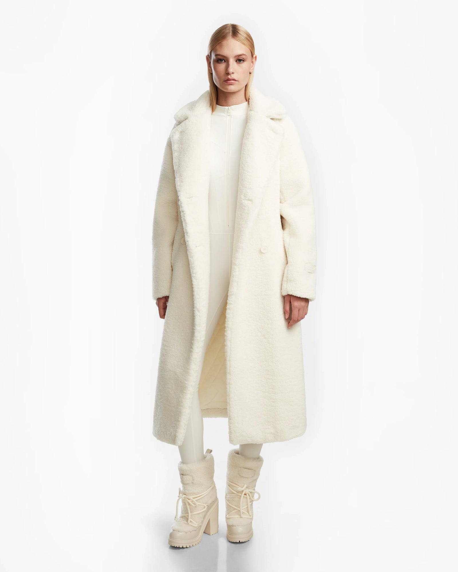 A woman with straight blonde hair and a neutral expression stands facing forward against a white background, wearing the Rudsak TINA - 8123578 Iceland long white trench coat over a white outfit and high-heeled lace-up boots.