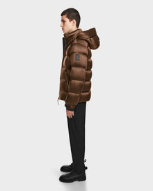 A person stands in profile wearing the Rudsak VINCE S - 6124560 Antique Bronze men’s quilted down puffer jacket with a hood, paired with black pants and boots, against a plain light background.