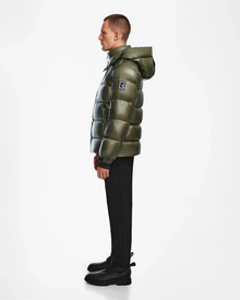 A person stands in profile wearing the Rudsak VINCE S - 6124560 Loden Green mens down puffer jacket with a hood, black pants, and black boots against a plain white background.