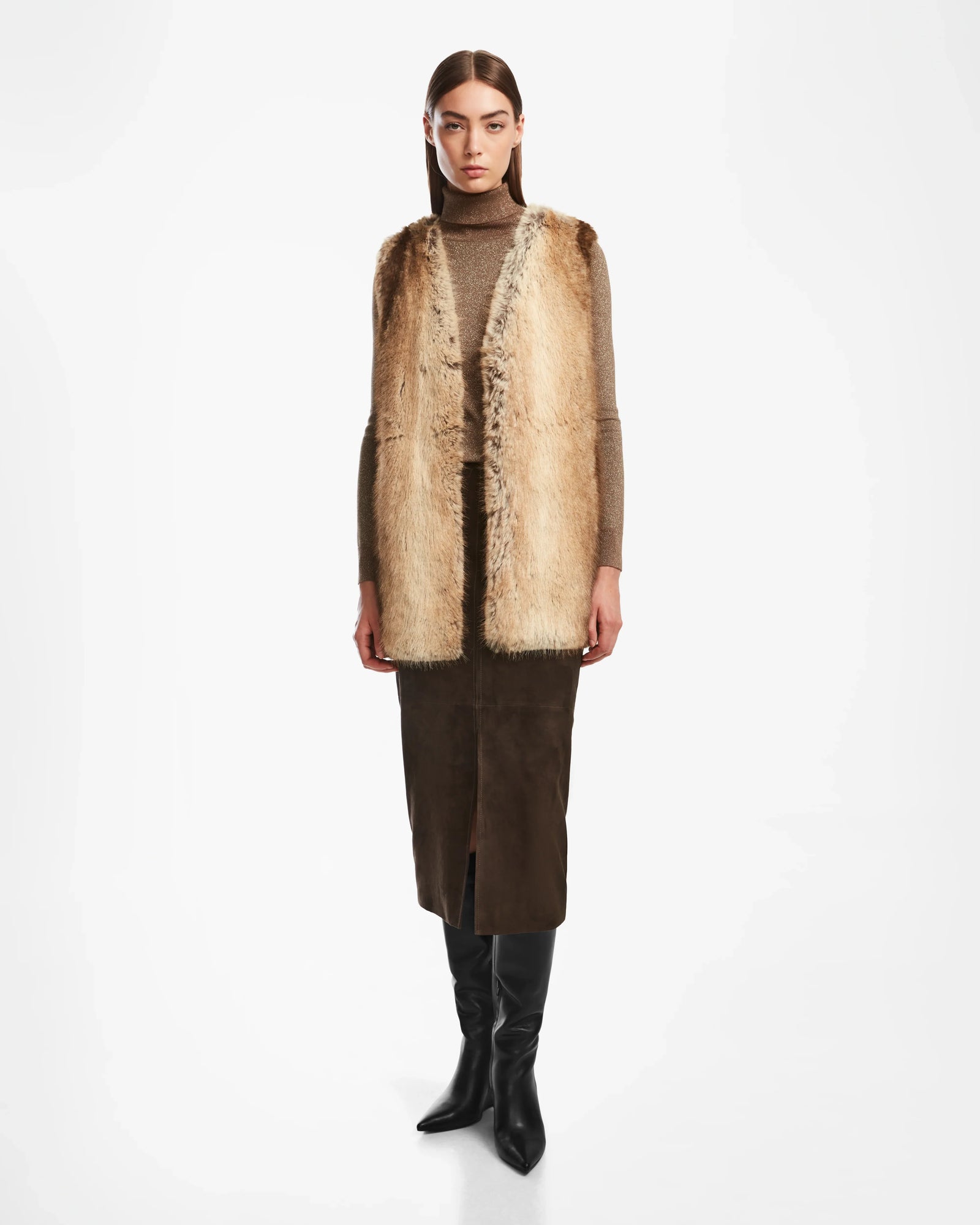 A woman faces forward in the Rudsak YOLANDA - 8124790 Mocha faux fur vest, layered over a brown turtleneck and dark brown skirt with a front slit, vegan leather belt, and black knee-high boots against a plain white background.