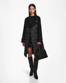A woman faces forward against a plain white background, wearing the Rudsak ZOE - 8124711 Black womens trench coat with a black dress, knee-high boots, and holding a large black handbag.