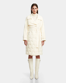 A woman with straight brown hair and a neutral expression stands facing forward, wearing the Rudsak ZOE - 8124711 Iceland cream quilted down trench with a wide collar, belted waist, and matching knee-high boots.
