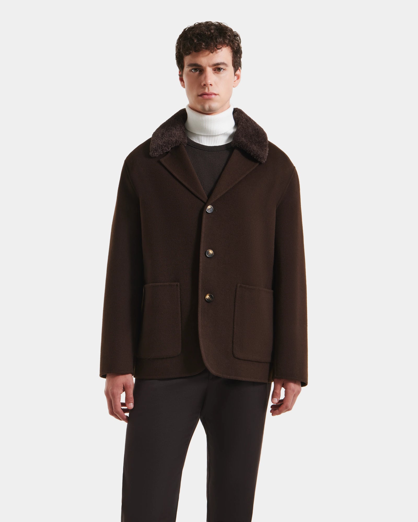 A man with short curly hair wears the Rudsak AIDEN - 6125558 Espresso wool-blend coat, featuring large buttons and front pockets, styled with dark pants and a white turtleneck against a white background.
