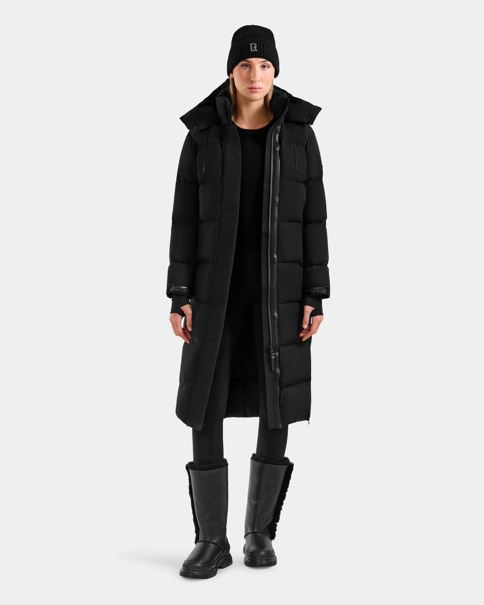 A person faces forward wearing the Rudsak ALESSANDRA L - L125553 Black quilted down puffer coat, black knit beanie, leggings, and boots—an effortlessly chic winter essential against a plain light background.