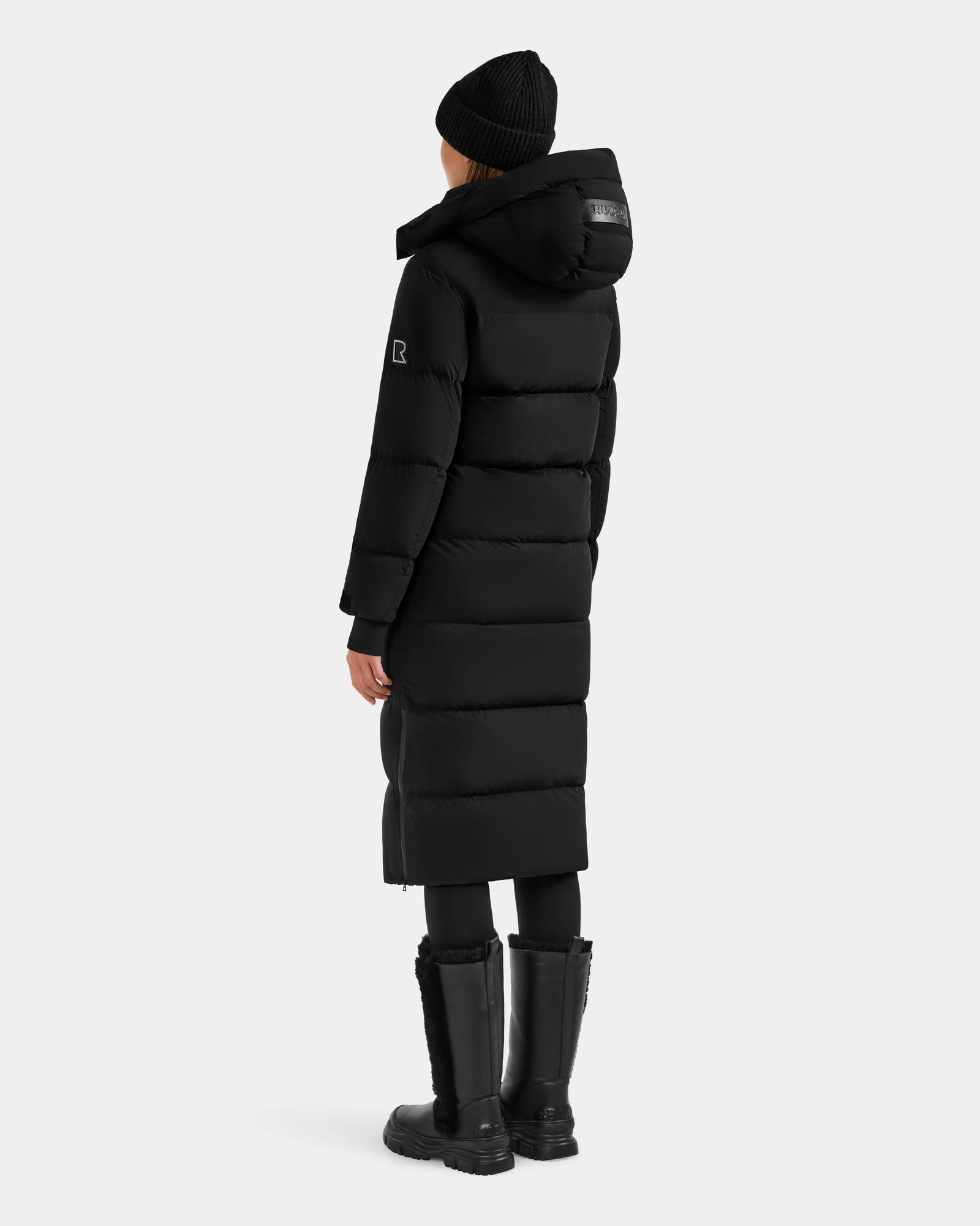 A person stands facing away against a plain white background wearing the Rudsak ALESSANDRA L - L125553 Black—a long quilted down puffer coat that's a true winter essential.
