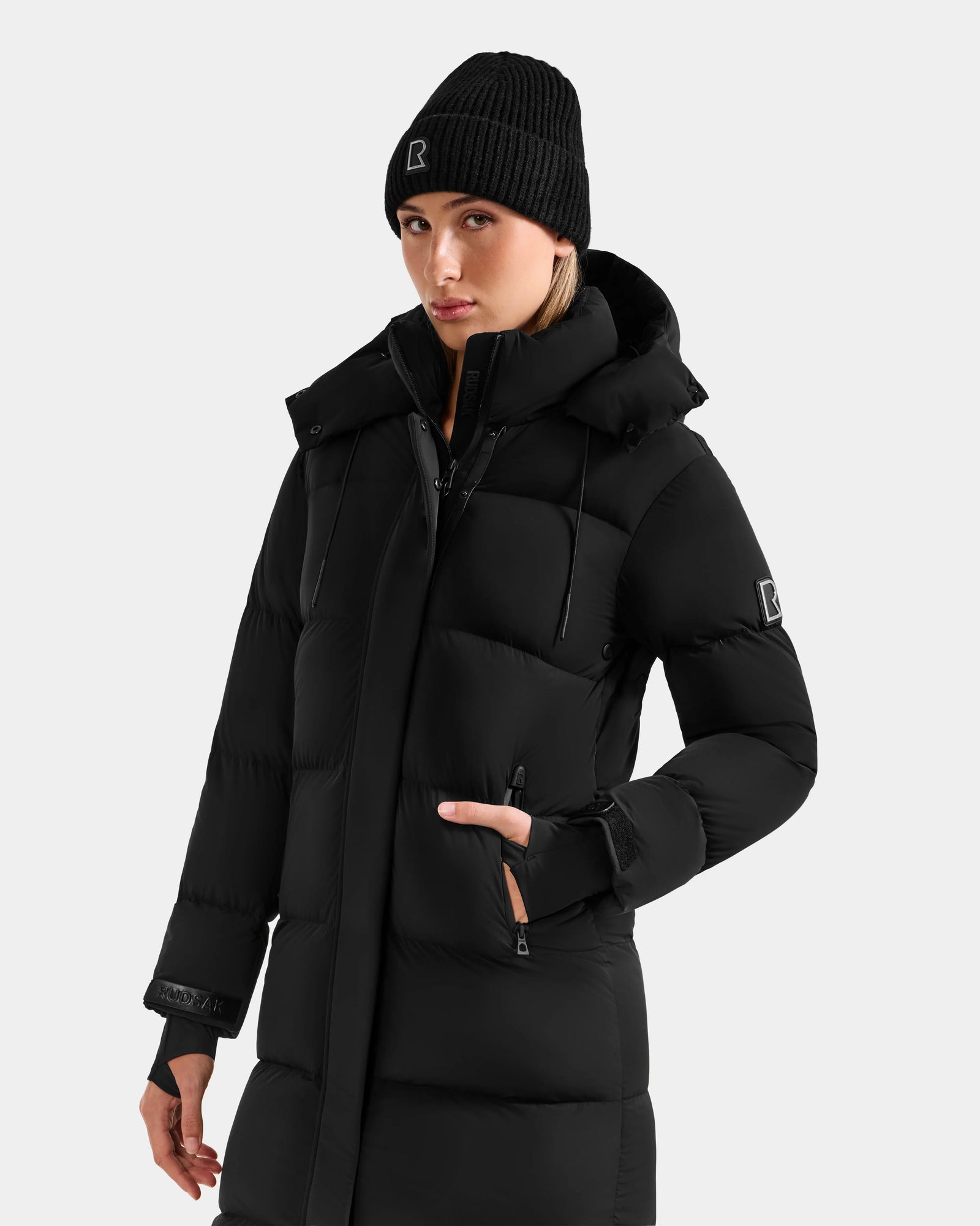 Wearing the Rudsak ALESSANDRA L - L125553 Black puffer with a high collar and a black knit beanie, one hand in pocket, this look against a plain light background perfectly captures winter essentials style.