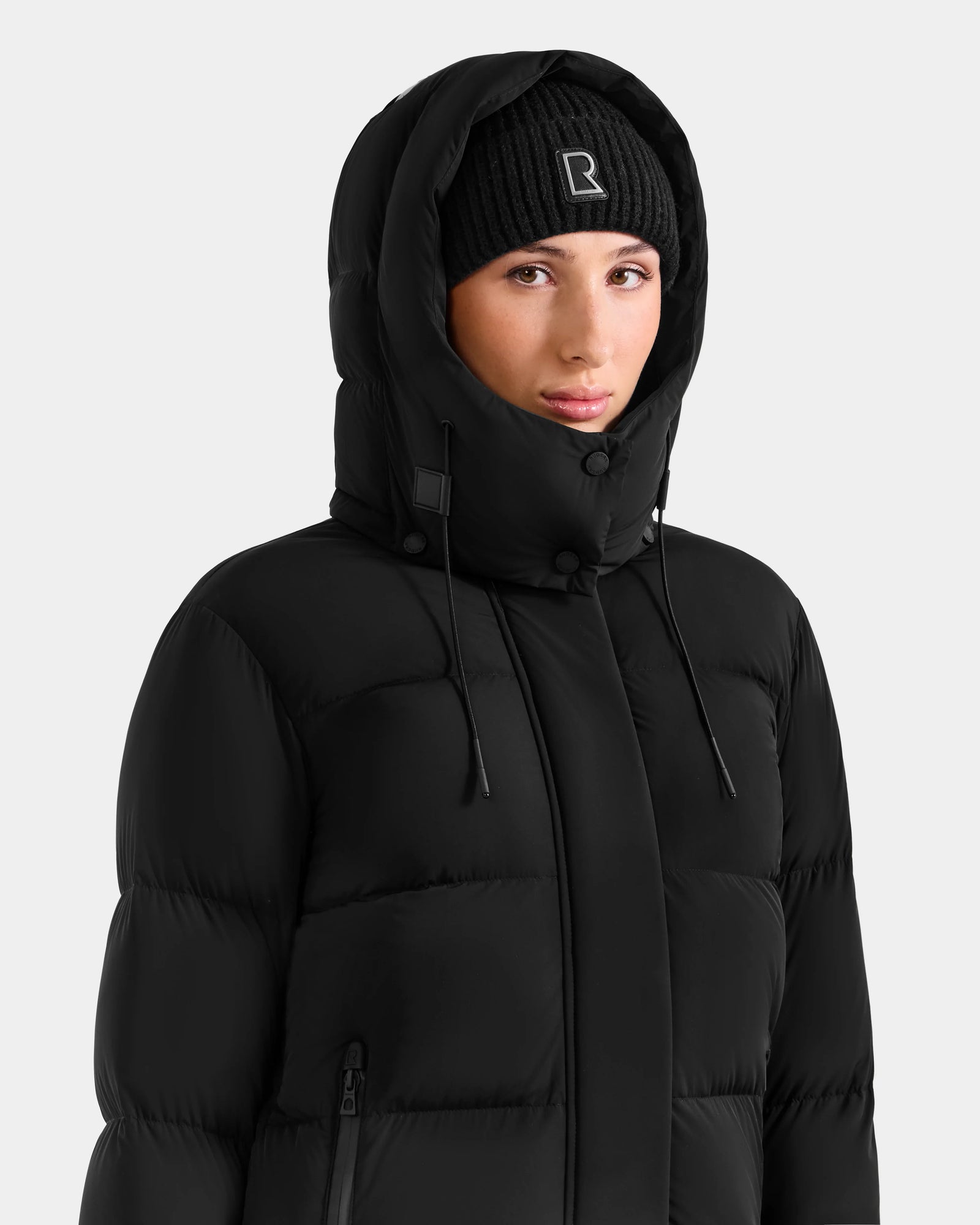Dressed in the Rudsak ALESSANDRA L - L125553 Black quilted down puffer jacket with the hood up and a black knit beanie with a stitched R logo—an understated winter essential against a light backdrop.