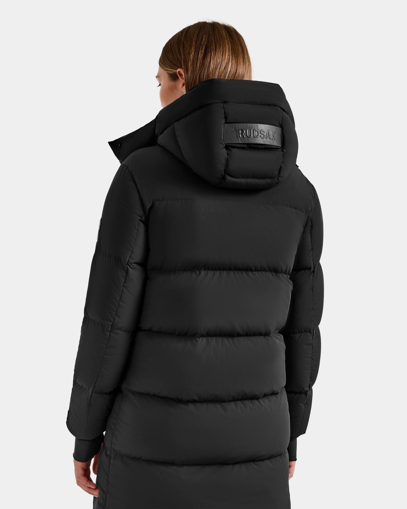 A person with straight, light brown hair is wearing the Rudsak ALESSANDRA M - L125552 Black women's long down puffer winter coat with a hood. The brand name "Rudsak" is on a rectangular patch at the back of the hood. The background is plain light gray.