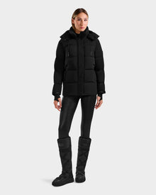 A woman with straight, shoulder-length hair and a neutral expression stands against a light gray background wearing the Rudsak ALESSANDRA S-M - L125551 Black down puffer jacket, paired with shiny black leggings and tall black winter boots.