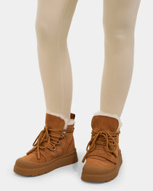 A person wears Rudsak’s ALEXEY - L225025 Tan women’s suede winter boots with cozy wool lining and rubber soles, paired with light beige leggings, standing on a plain light background.