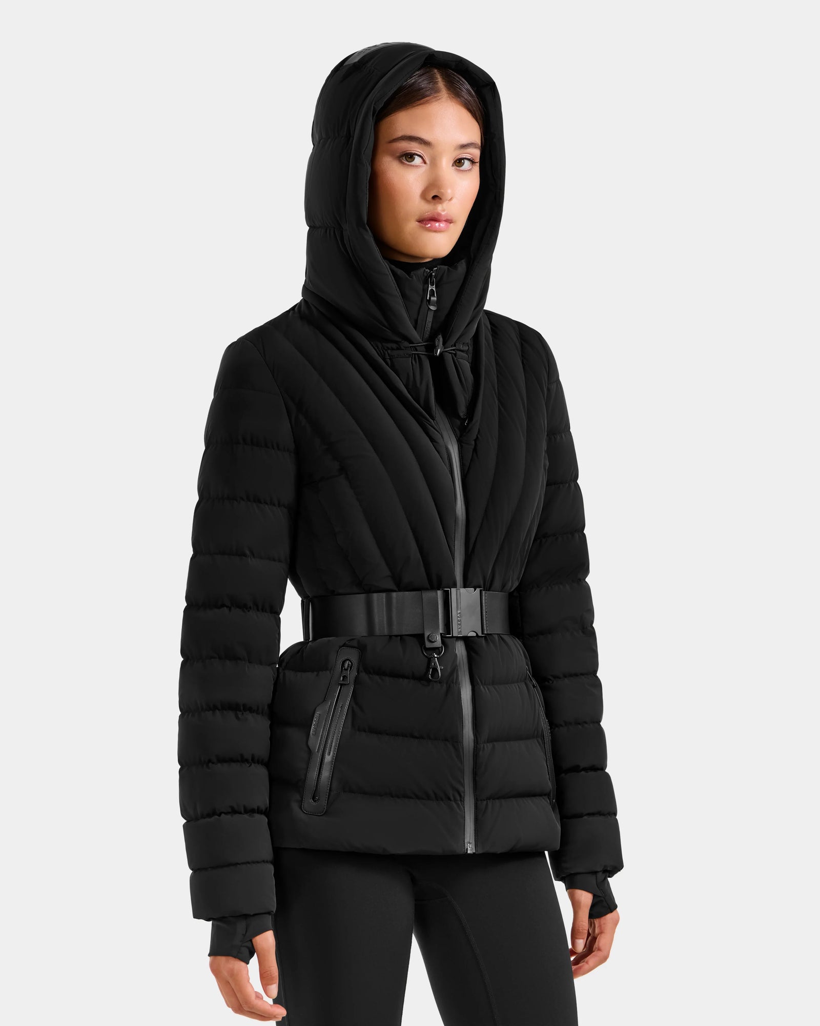 A woman wears the Rudsak ALISA F - L125527 Black belted, hooded quilted puffer jacket with zippered pockets, standing against a plain light gray background.