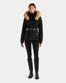 A woman faces forward wearing the Rudsak ALISA F - L125527 Black quilted puffer jacket with a faux-fur trim hood, paired with black leggings and boots, set against a plain light gray background.