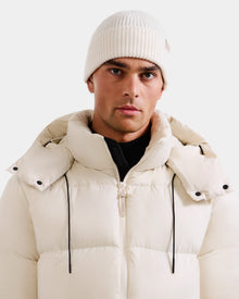 A man wears Rudsak's ANDREY - U425507 Vanilla white puffer jacket with a hood and a wool-cashmere beanie, looking directly at the camera against a plain light background.