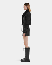 A woman in profile models the Rudsak ARIELLA - 8125505 Black down-filled jacket with a black mini skirt and knee-high drawstring boots, showcasing effortless cold-weather style against a plain light background.