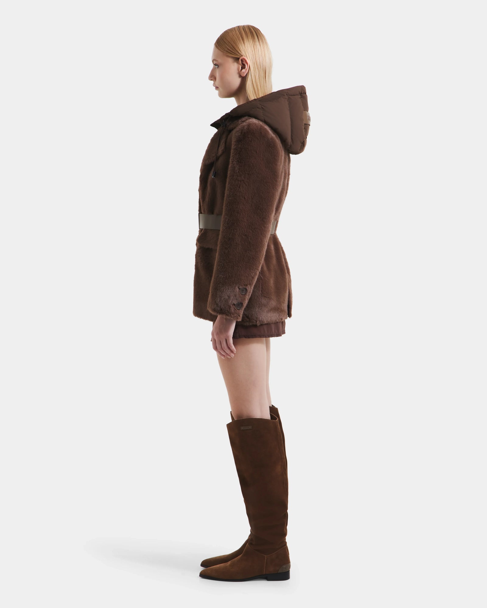 A woman with straight blonde hair faces left, modeling the Rudsak ASPEN F - 8125604 Mocha faux fur jacket with a removable down-insulated hood and tall brown suede boots, standing against a plain white background.