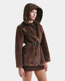 A woman models the Rudsak ASPEN F - 8125604 Mocha faux fur jacket with removable hooded dickie and wide belt over a brown mini skirt, posing against a plain white background.