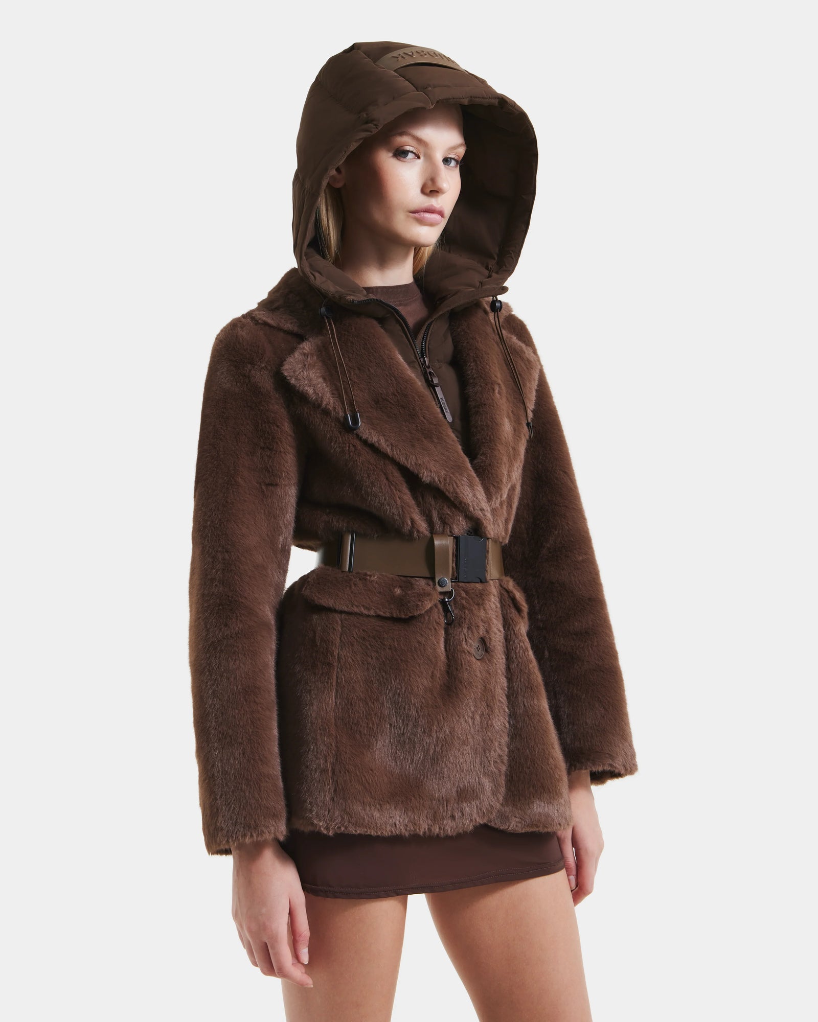 A woman models the Rudsak ASPEN F - 8125604 Mocha faux fur jacket with removable hooded dickie and wide belt over a brown mini skirt, posing against a plain white background.