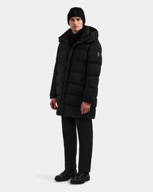 A person showcases winter versatility wearing the Rudsak BRAYDEN M-M - M125535 Black men’s down puffer coat, styled with black pants, a black knit beanie, and black shoes against a plain white background.