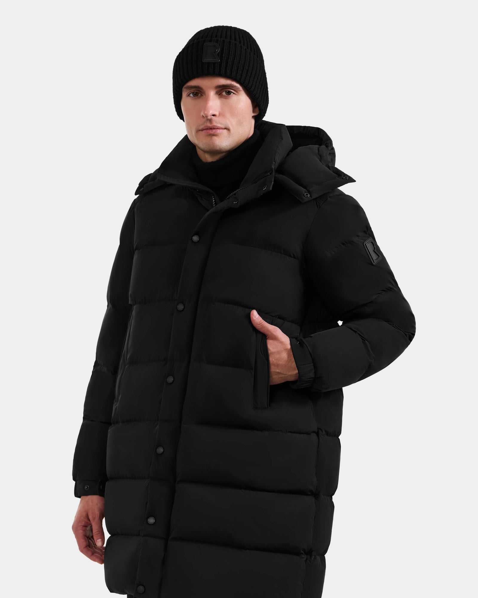 A person models the Rudsak BRAYDEN M-M - M125535 Black men’s down puffer coat with hood, paired with a black beanie, standing against a light gray background and highlighting its winter versatility.