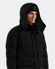 A man wearing a Rudsak BRAYDEN M-M - M125535 Black down puffer jacket with a hood and a black knit beanie stands against a plain light background.