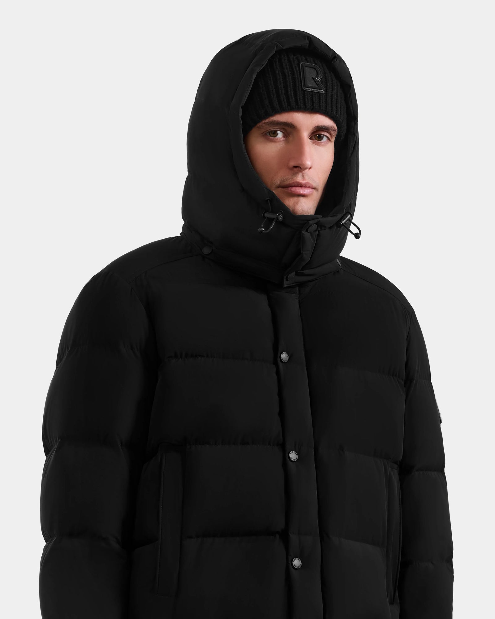 A man wears the Rudsak BRAYDEN M-M - M125535 Black down puffer jacket with the hood up and a black beanie, looking forward against a plain light gray background, ready for winter versatility.
