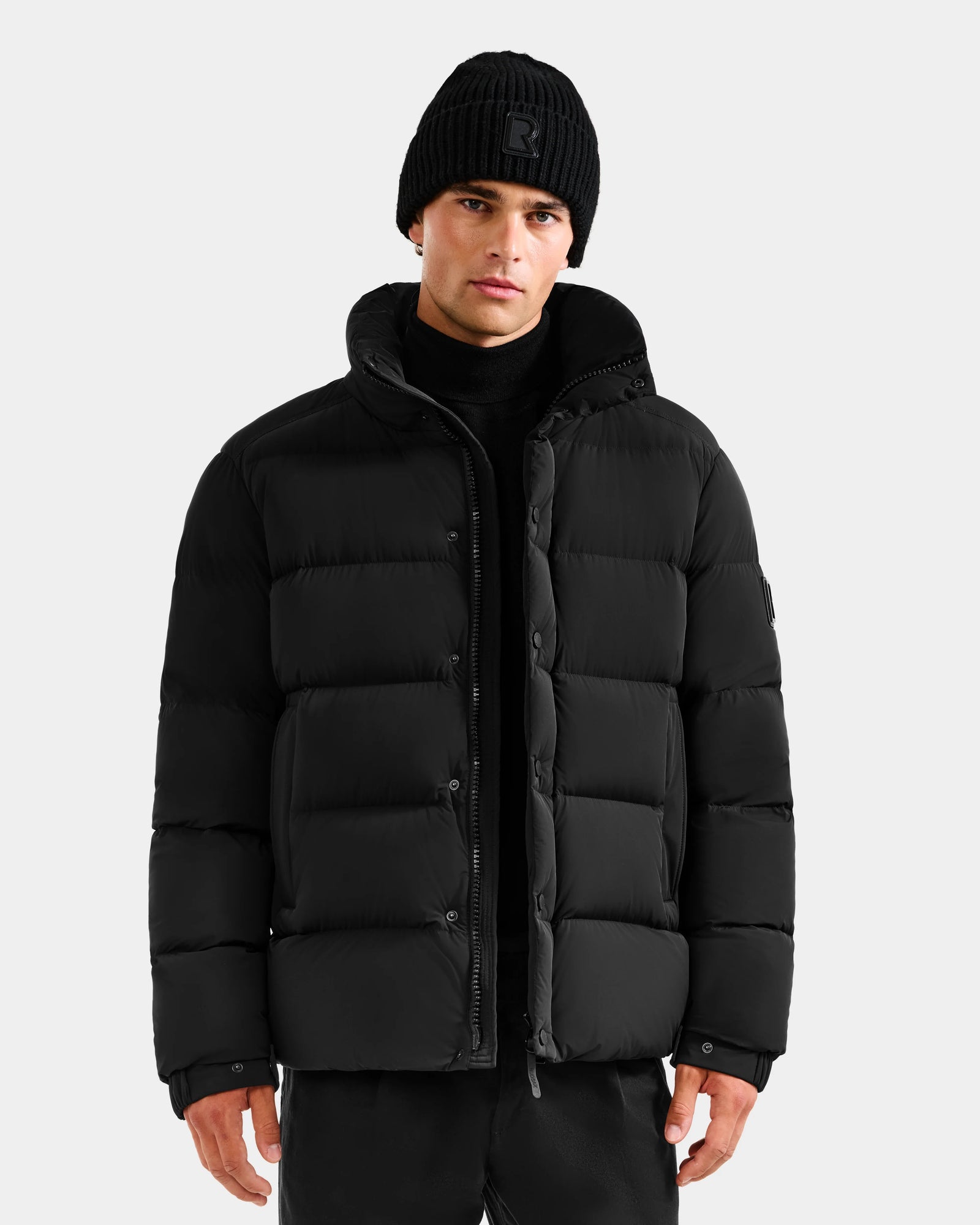 A man stands facing forward against a light gray background, wearing the Rudsak BRAYDEN M-S - M125524 Black down puffer jacket, paired with a black beanie and black pants.