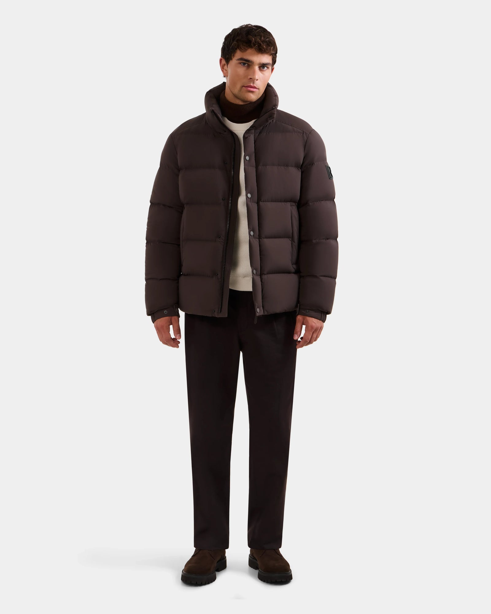 A man faces forward wearing Rudsak’s BRAYDEN M-S - M125524 Espresso men’s down puffer jacket with RDS-certified down, layered over a white sweater, dark trousers, and dark shoes against a plain light background.