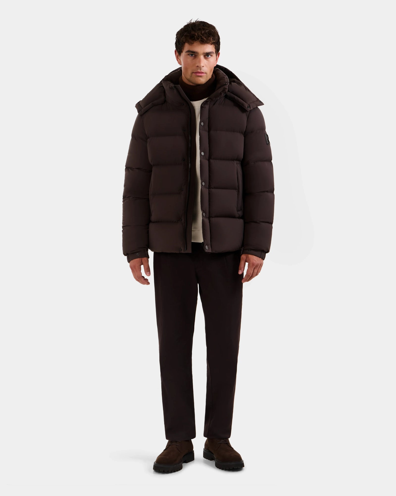 A man faces forward wearing the Rudsak BRAYDEN M-S - M125524 Espresso men's down puffer jacket, dark pants, scarf, and brown shoes, standing against a plain white background.