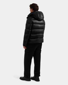 A person faces away, wearing the Rudsak BRAYDEN S - M125525 Black men's quilted down puffer jacket with hood, paired with black pants and shoes against a plain light background.