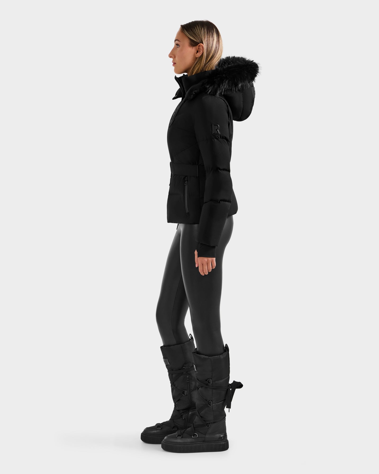 A woman in profile wears the Rudsak CHRISTELLE M - L125542 Black weather-resistant hooded puffer jacket with faux fur trim, black leggings, and tall black boots against a plain light gray background.