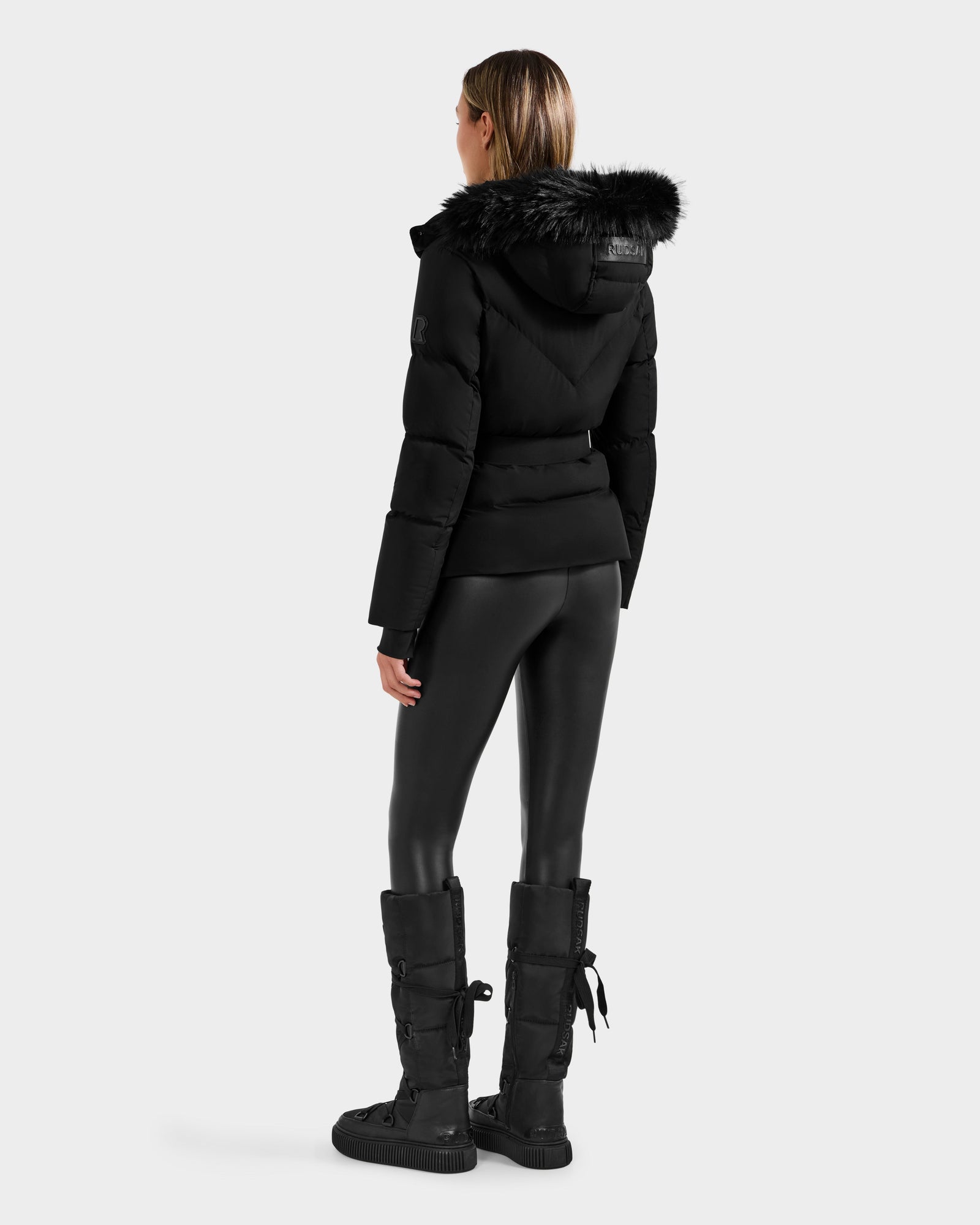 A woman faces away, wearing the Rudsak CHRISTELLE M - L125542 Black weather-resistant jacket with faux fur trim, paired with black leggings and tall lace-up, buckled boots against a plain light background.