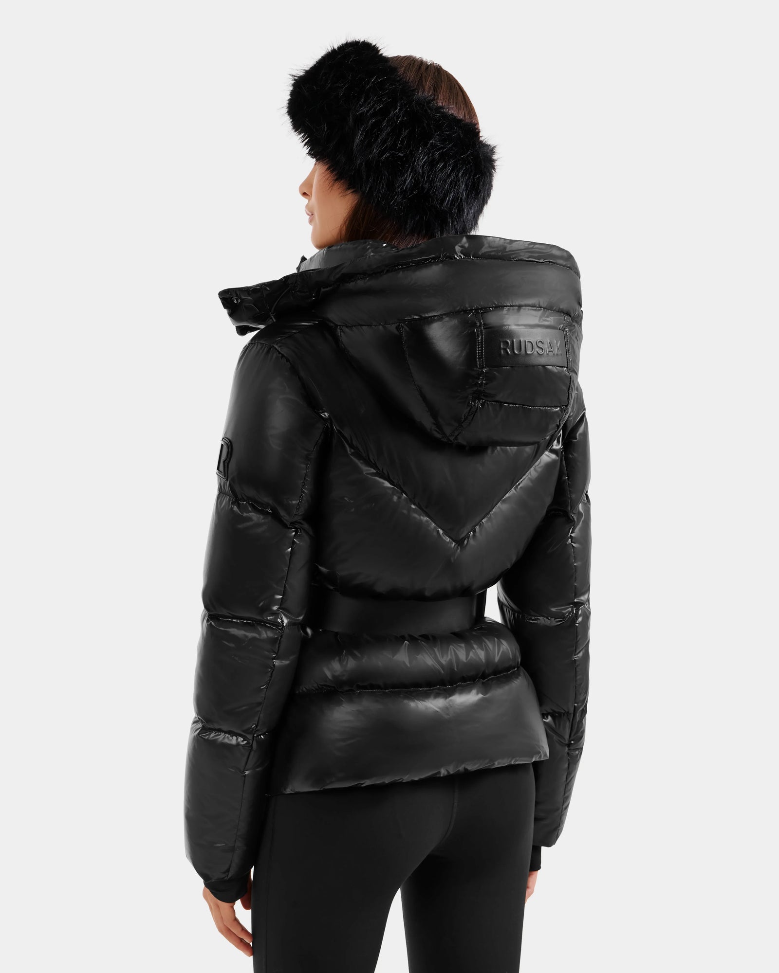 A woman is seen from the back wearing the Rudsak CHRISTELLE S - L125544 Black shiny down puffer jacket with a belted waist and faux fur trim headband, set against a plain light background.